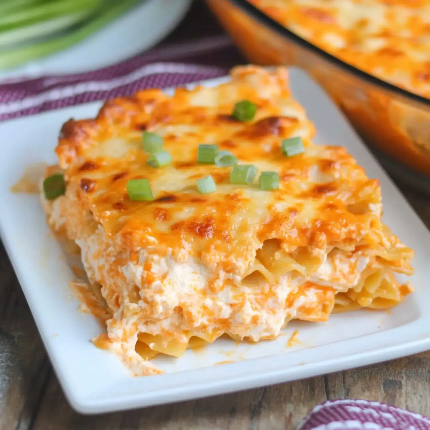 A portion of creamy layered pasta garnished with green onions on a white dish, with the main baking dish showing in the background.