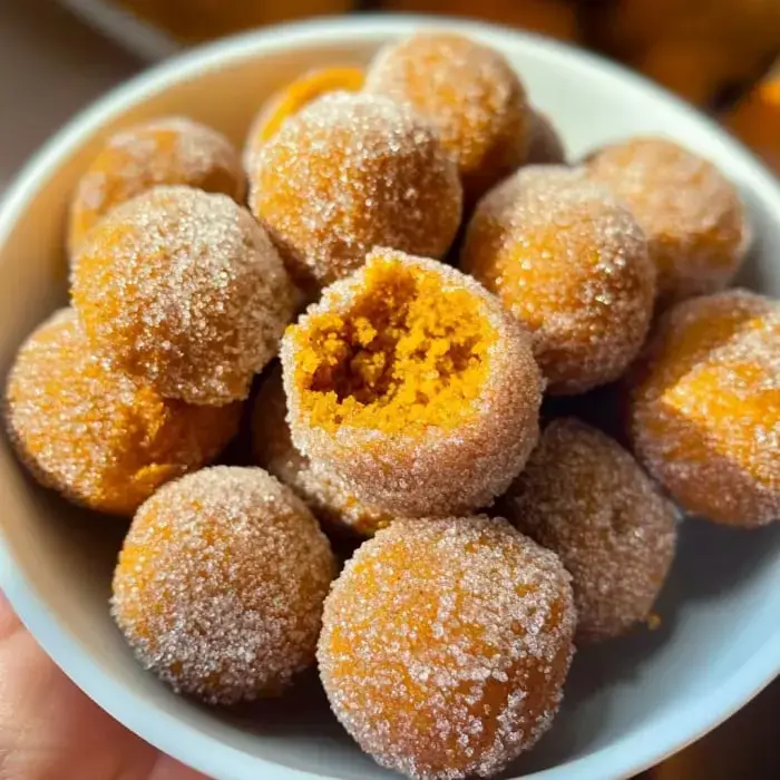 A bowl of sugar-topped orange donuts with one showing its soft center.