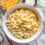 A bowl of creamy macaroni and cheese with shredded cheese and grated parmesan in the background.